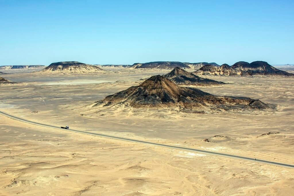Vista aérea del Desierto Negro de Bahariya con monticulos volcanicos oscuros sobre arena pálida, una carretera recta y cielo azul intenso