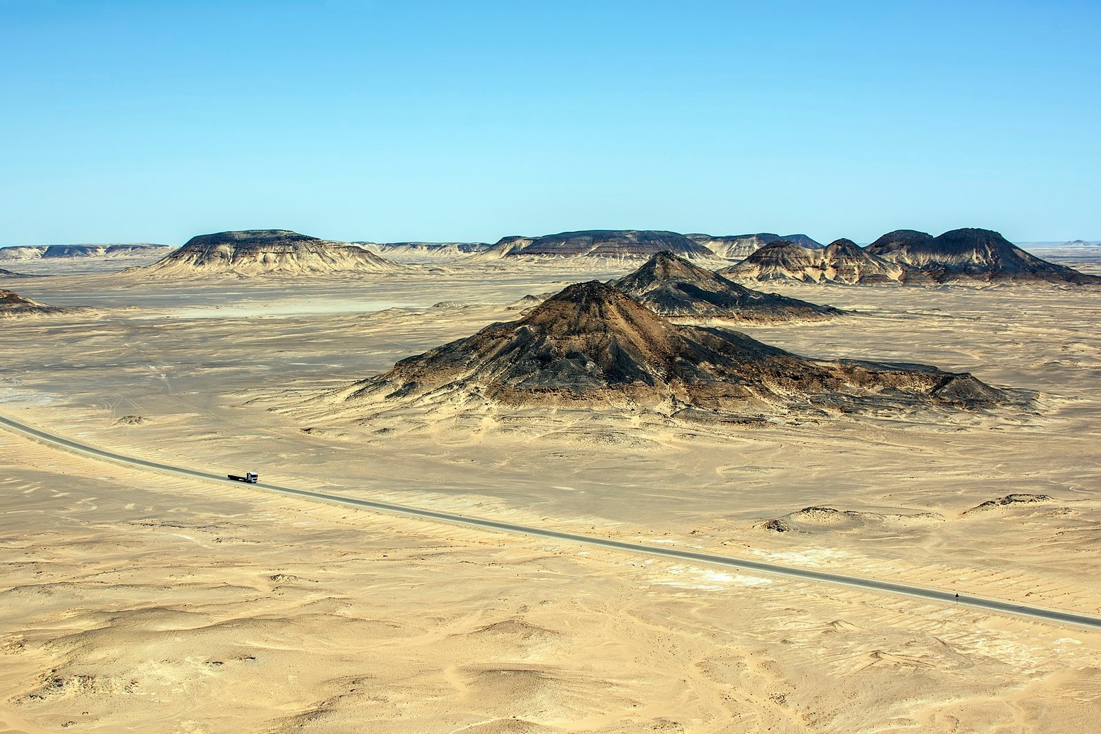 Vista aerea del Desierto Negro de Bahariya con monticulos volcanicos oscuros sobre arena palida, una carretera recta y cielo azul intenso
