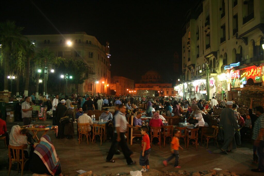 Plaza nocturna animada de Jan el-Jalili en El Cairo con multitudes en cafes al aire libre, palmeras iluminadas y la mezquita historica al fondo, Egipto para mochileros