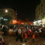 Plaza nocturna animada de Jan el-Jalili en El Cairo con multitudes en cafes al aire libre, palmeras iluminadas y la mezquita historica al fondo, Egipto para mochileros