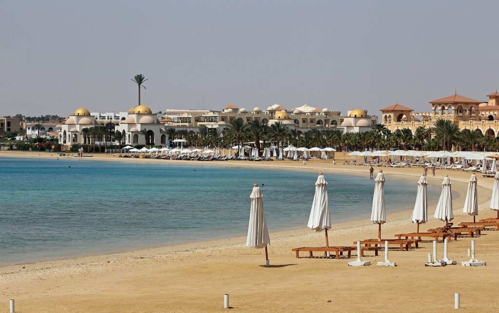 Playa de resort de lujo en el Mar Rojo de Egipto con arena dorada, agua turquesa, sombrillas blancas alineadas y arquitectura de cupulas doradas al fondo en verano