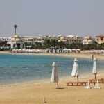 Playa de resort de lujo en el Mar Rojo de Egipto con arena dorada, agua turquesa, sombrillas blancas alineadas y arquitectura de cupulas doradas al fondo en verano