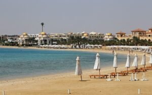 Playa de resort de lujo en el Mar Rojo de Egipto con arena dorada, agua turquesa, sombrillas blancas alineadas y arquitectura de cupulas doradas al fondo en verano