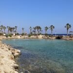 Cala de aguas turquesas y cristalinas del Mar Rojo en Egipto, bordeada de rocas doradas, palmeras y sombrillas de paja bajo un cielo azul intenso