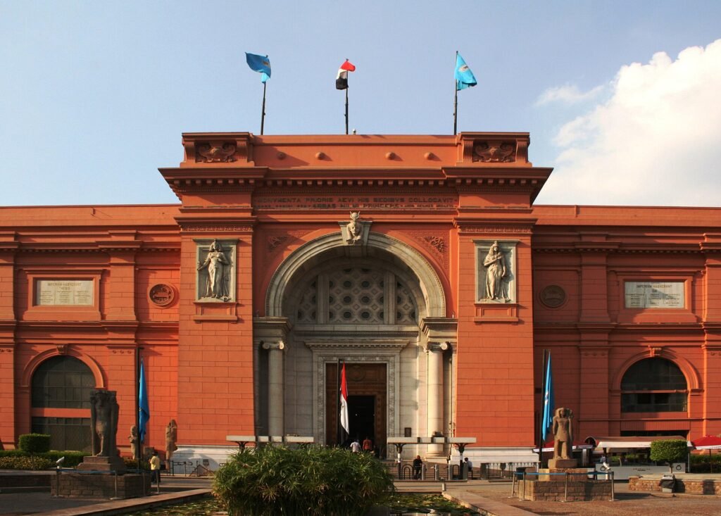 Fachada frontal del Museo Egipcio de Tahrir en El Cairo, edificio neoclasico de color rosa salmon iluminado por luz dorada, con su gran arco central, banderas y cielo azul