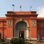 Fachada frontal del Museo Egipcio de Tahrir en El Cairo, edificio neoclasico de color rosa salmon iluminado por luz dorada, con su gran arco central, banderas y cielo azul
