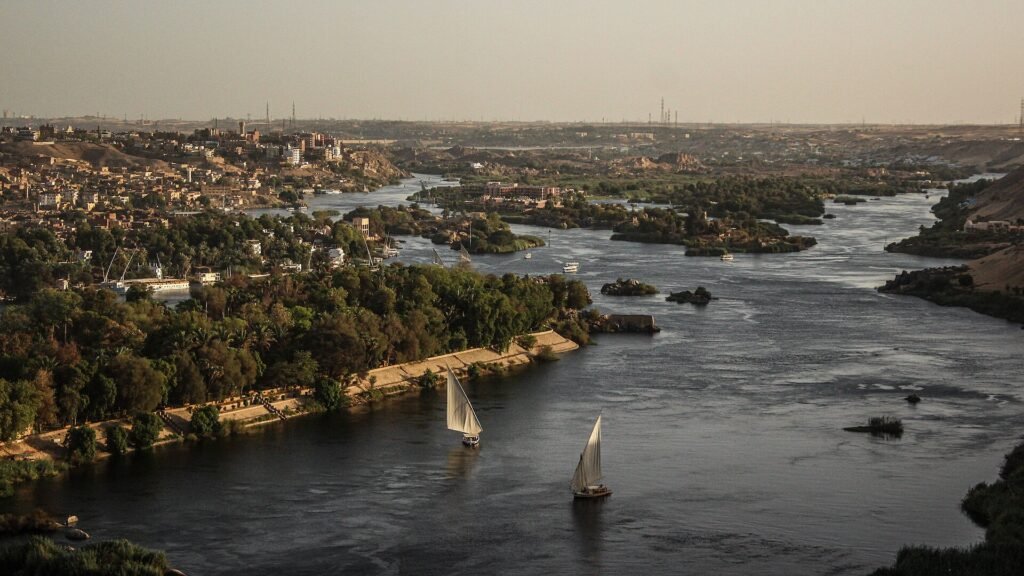 Vista aerea del rio Nilo en Asuan a la luz dorada del atardecer con dos feluccas de vela blanca navegando entre islas verdes y la ciudad en la orilla