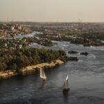 Vista panoramica del Nilo en Asuan al atardecer con dos feluccas de velas blancas navegando entre islas verdes y la ciudad al fondo