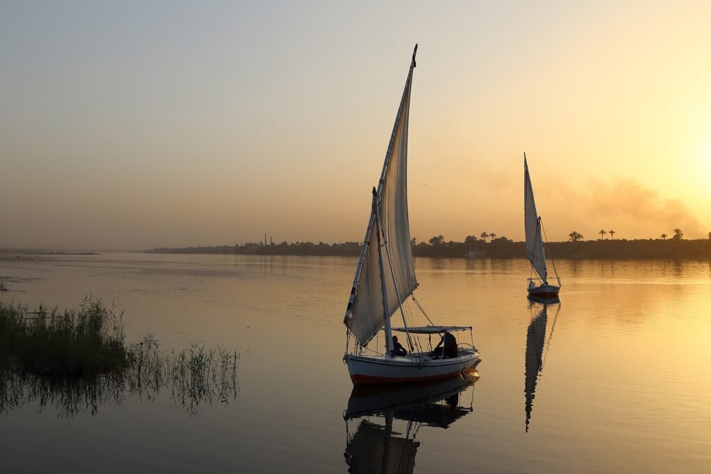 Dos feluconas de vela blanca navegando sobre las aguas serenas y doradas del rio Nilo al atardecer en Egipto con reflejos en el agua
