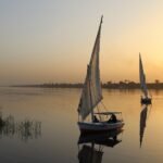 Dos feluconas de vela blanca navegando sobre las aguas serenas y doradas del rio Nilo al atardecer en Egipto con reflejos en el agua