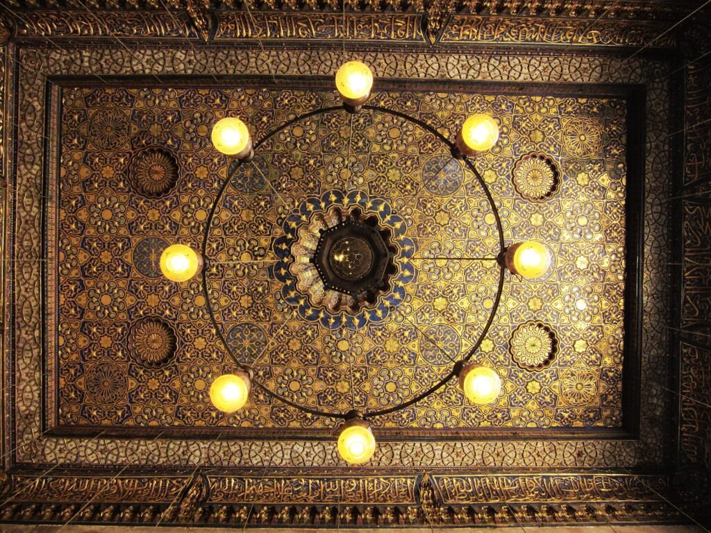 Looking straight up at a gilded and painted wooden ceiling in an Ottoman era Cairo interior, dense geometric panels with a ring of glass lamps and an Arabic inscription band. Unrelated to costs