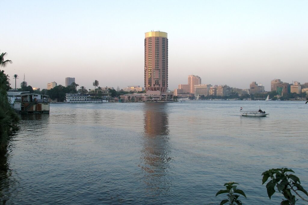The Nile at Cairo at dusk with a round tower hotel block on the far bank, the Cairo Tower to the left and a small flagged boat crossing the water