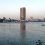 The Nile at Cairo at dusk with a round tower hotel block on the far bank, the Cairo Tower to the left and a small flagged boat crossing the water