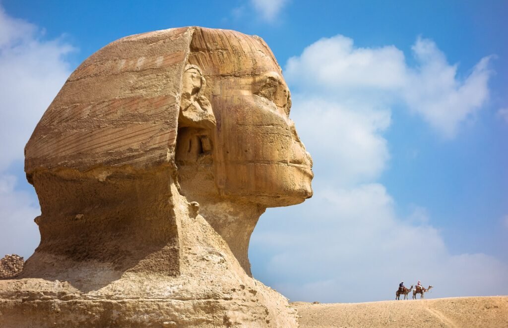 The head of the Great Sphinx of Giza seen in close profile from the left, the eroded limestone nemes headdress and face filling the frame against a blue sky with white cloud, and two camel riders crossing the sand ridge far behind it.