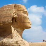 The head of the Great Sphinx of Giza seen in close profile from the left, the eroded limestone nemes headdress and face filling the frame against a blue sky with white cloud, and two camel riders crossing the sand ridge far behind it.