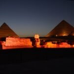 La Esfinge y dos piramides de Giza con iluminacion calida dorada durante el espectaculo nocturno de luz y sonido, recortadas contra un cielo azul profundo al anochecer