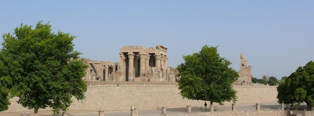 Templo de Kom Ombo a orillas del Nilo en Egipto, con sus columnas talladas enmarcadas por arboles verdes y cielo calido y despejado
