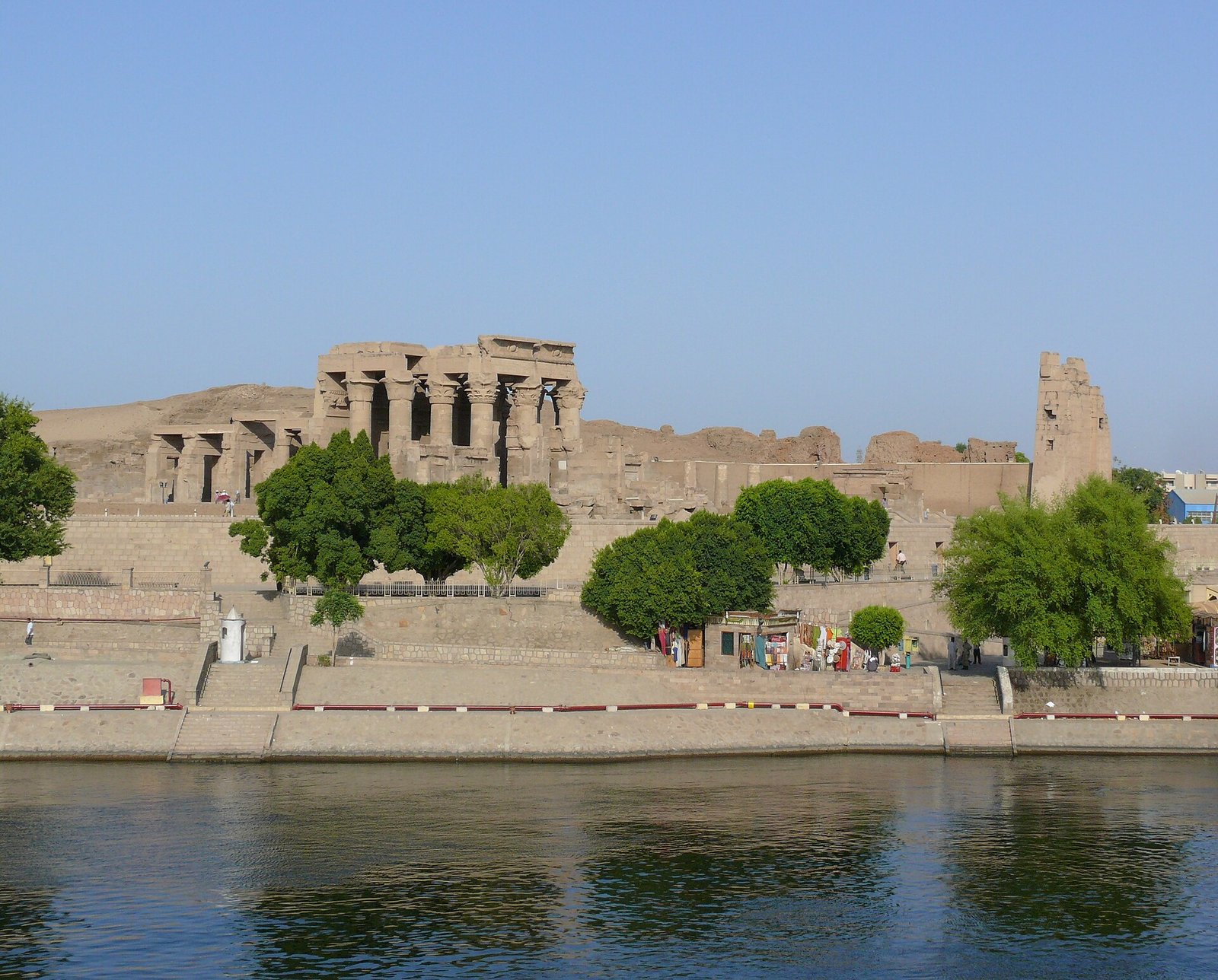 Vista del templo de Kom Ombo desde el rio Nilo en Egipto, con el santuario sobre la ribera, palmeras y agua tranquila en primer plano