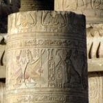Close-up of a carved sandstone temple column still holding traces of its original paint, with bands of hieroglyphs, a winged goddess figure and a falcon-headed deity beside a royal cartouche, lit by low warm sun with further inscribed columns behind; the Ptolemaic style and surviving colour match the temples of the Upper Egyptian Nile bank such as Kom Ombo.