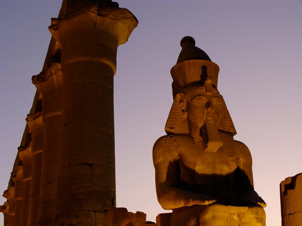 Coloso monumental de Ramses II en el templo de Luxor iluminado por la luz dorada del atardecer junto a una gran columna de papiro bajo cielo azul