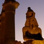 Coloso monumental de Ramses II en el templo de Luxor iluminado por la luz dorada del atardecer junto a una gran columna de papiro bajo cielo azul