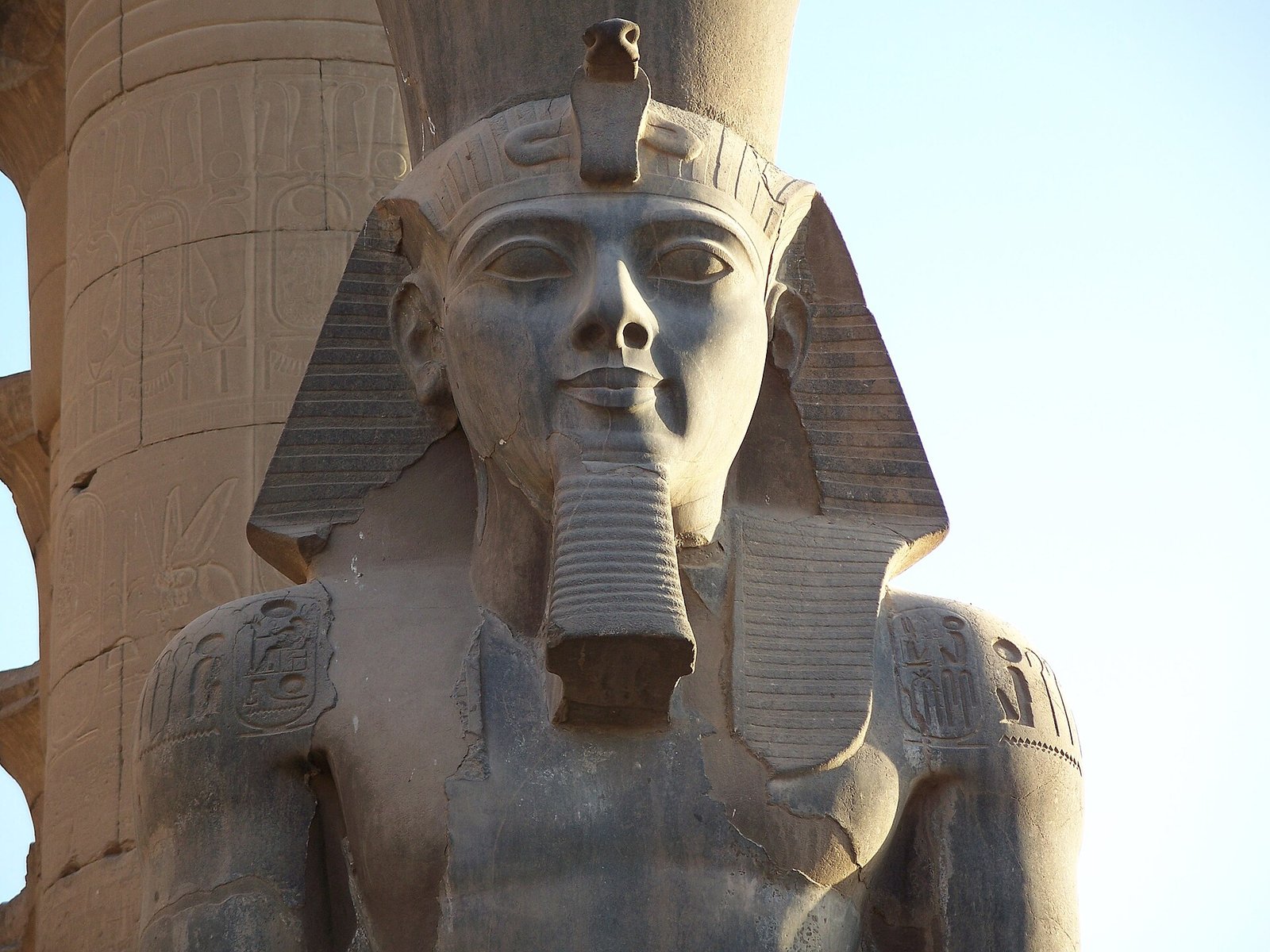 Primer plano del rostro tallado de la colosal estatua de Ramses II en Luxor con la cobra real y la barba ceremonial bajo un cielo azul claro