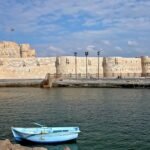 La fortaleza de Qaitbay en Alejandria, Egipto, vista desde el agua con un pequeno bote azul y el mar Mediterraneo en primer plano bajo cielo despejado