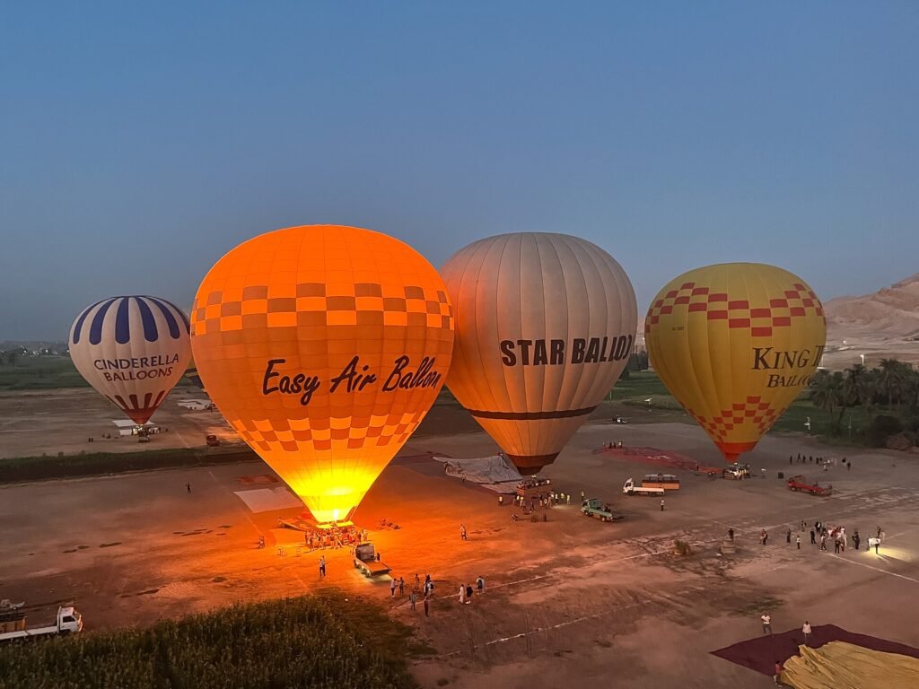Globos aerostaticos coloridos naranja, blanco y amarillo inflandose al alba en el campo de despegue de Luxor con el resplandor de los quemadores