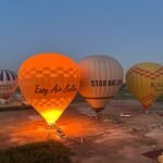Globos aerostaticos coloridos naranja, blanco y amarillo inflandose al alba en el campo de despegue de Luxor con el resplandor de los quemadores