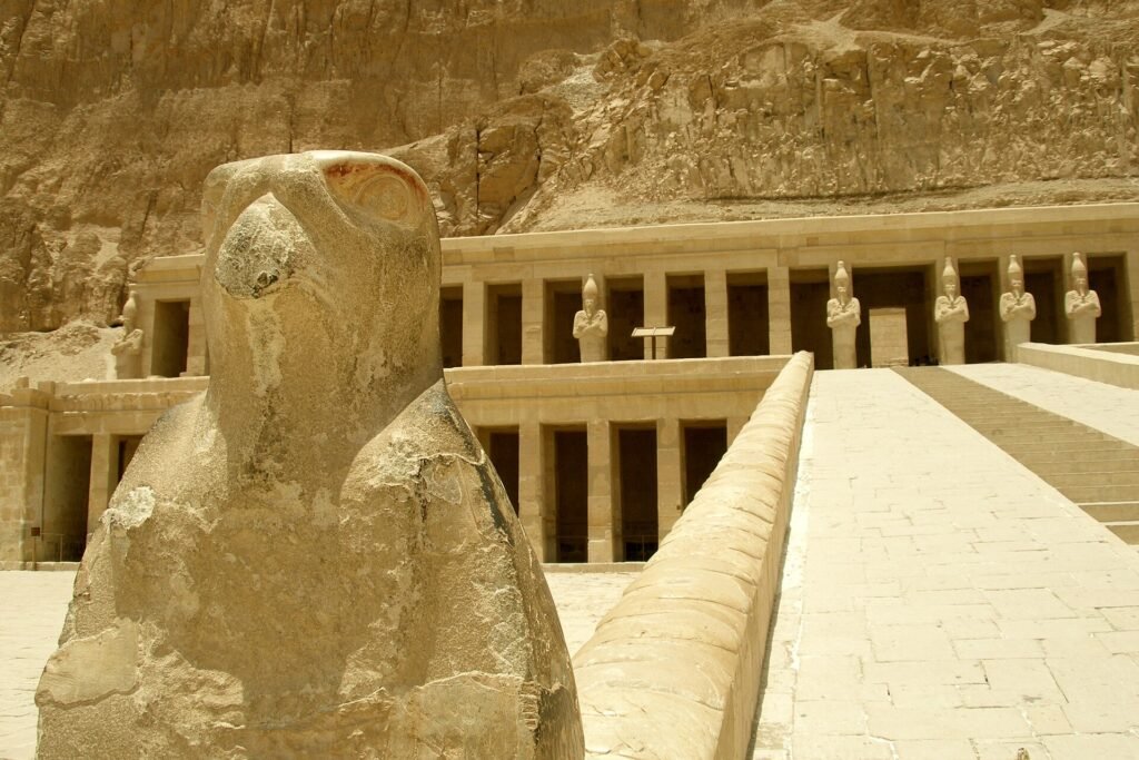 Estatua de halcon Horus en piedra dorada en primer plano con las terrazas y columnatas del templo de Hatshepsut en Deir el-Bahari al fondo bajo cielo claro