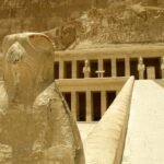 Estatua de halcon Horus en piedra dorada en primer plano con las terrazas y columnatas del templo de Hatshepsut en Deir el-Bahari al fondo bajo cielo claro