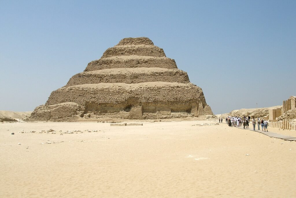 Vista frontal y limpia de la Piramide Escalonada de Zoser en Saqqara, obra maestra de Imhotep, con sus seis niveles de piedra recortados contra un cielo azul intenso sobre la arena del desierto