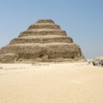 Vista frontal y limpia de la Piramide Escalonada de Zoser en Saqqara, obra maestra de Imhotep, con sus seis niveles de piedra recortados contra un cielo azul intenso sobre la arena del desierto