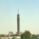 Torre de El Cairo iluminada por el sol sobre el horizonte de la ciudad con palmeras y cielo azul claro, vista urbana premium de Egipto