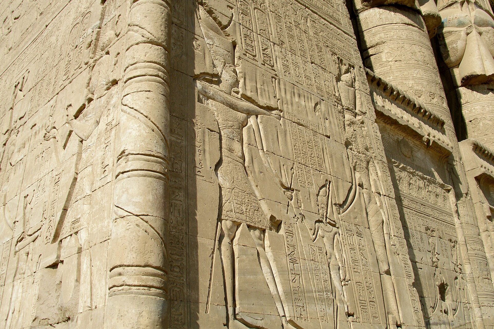 Relieve tallado en el muro exterior de un templo egipcio con grandes figuras y columnas de jeroglificos grabados en piedra dorada bajo luz solar