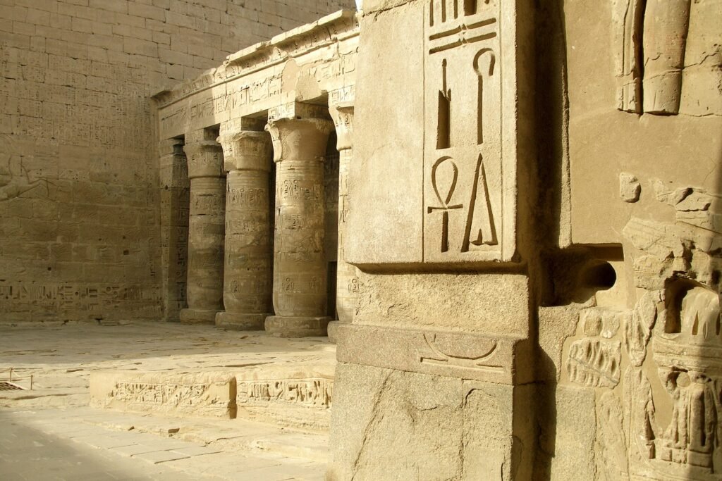 Muro de templo egipcio en piedra caliza con jeroglíficos tallados, incluido el simbolo ankh, junto a una columnata bajo la luz dorada del sol