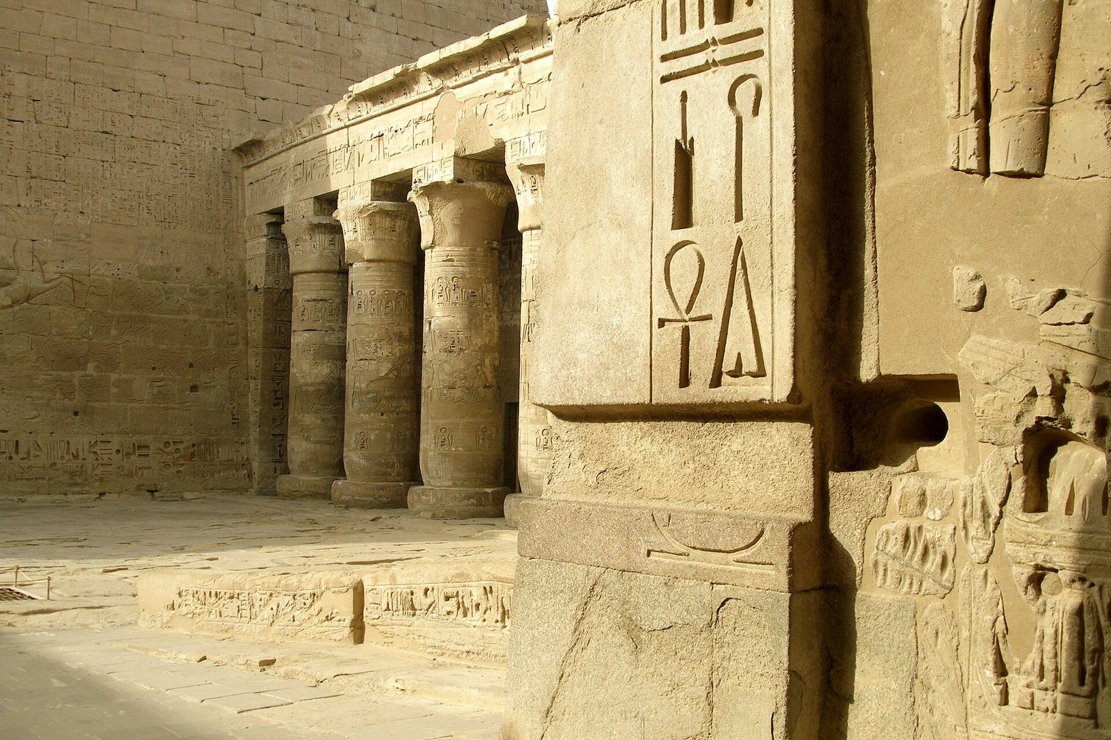 Muro de templo egipcio en piedra caliza con jeroglificos tallados, incluido el simbolo ankh, junto a una columnata bajo la luz dorada del sol