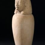 Vaso canopo egipcio de alabastro con tapa tallada en forma de cabeza humana sobre fondo oscuro, pieza funeraria del antiguo proceso de momificacion