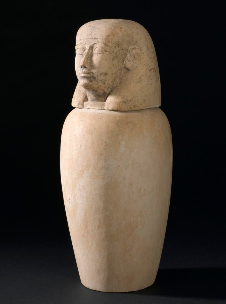 Vaso canopo egipcio de alabastro con tapa tallada en forma de cabeza humana sobre fondo oscuro, pieza funeraria del antiguo proceso de momificacion