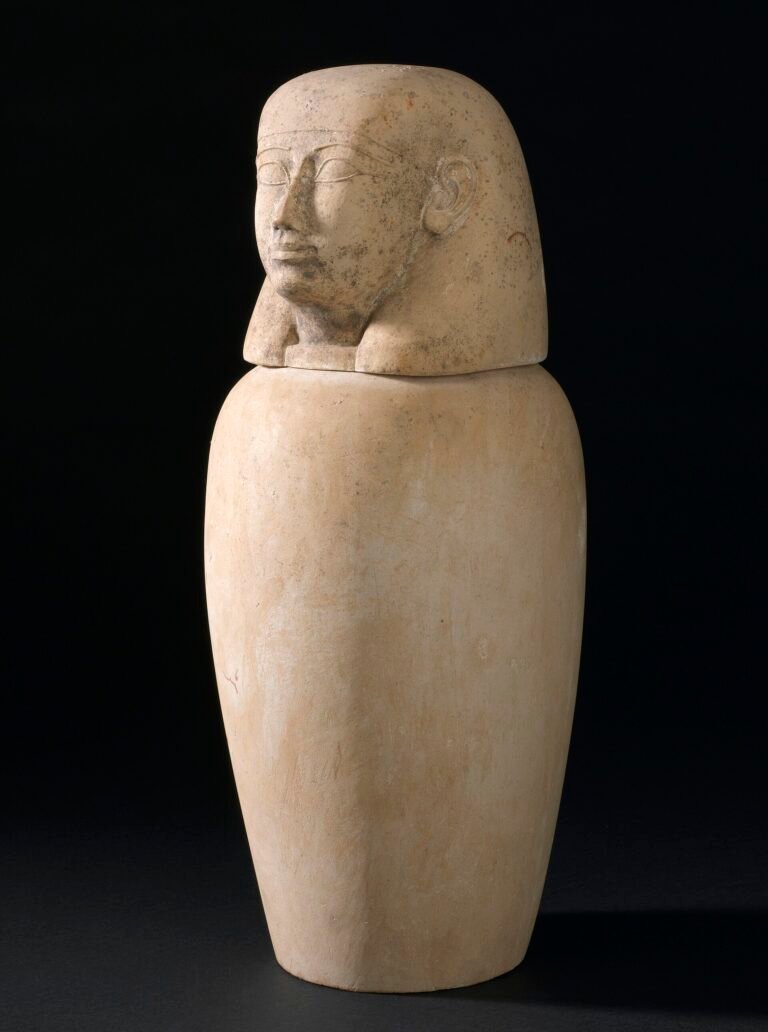 Vaso canopo egipcio de alabastro con tapa tallada en forma de cabeza humana sobre fondo oscuro, pieza funeraria del antiguo proceso de momificacion