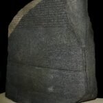La Piedra de Rosetta de granodiorita oscura sobre pedestal claro y fondo negro, con sus tres escrituras jeroglifica, demotica y griega nitidamente talladas