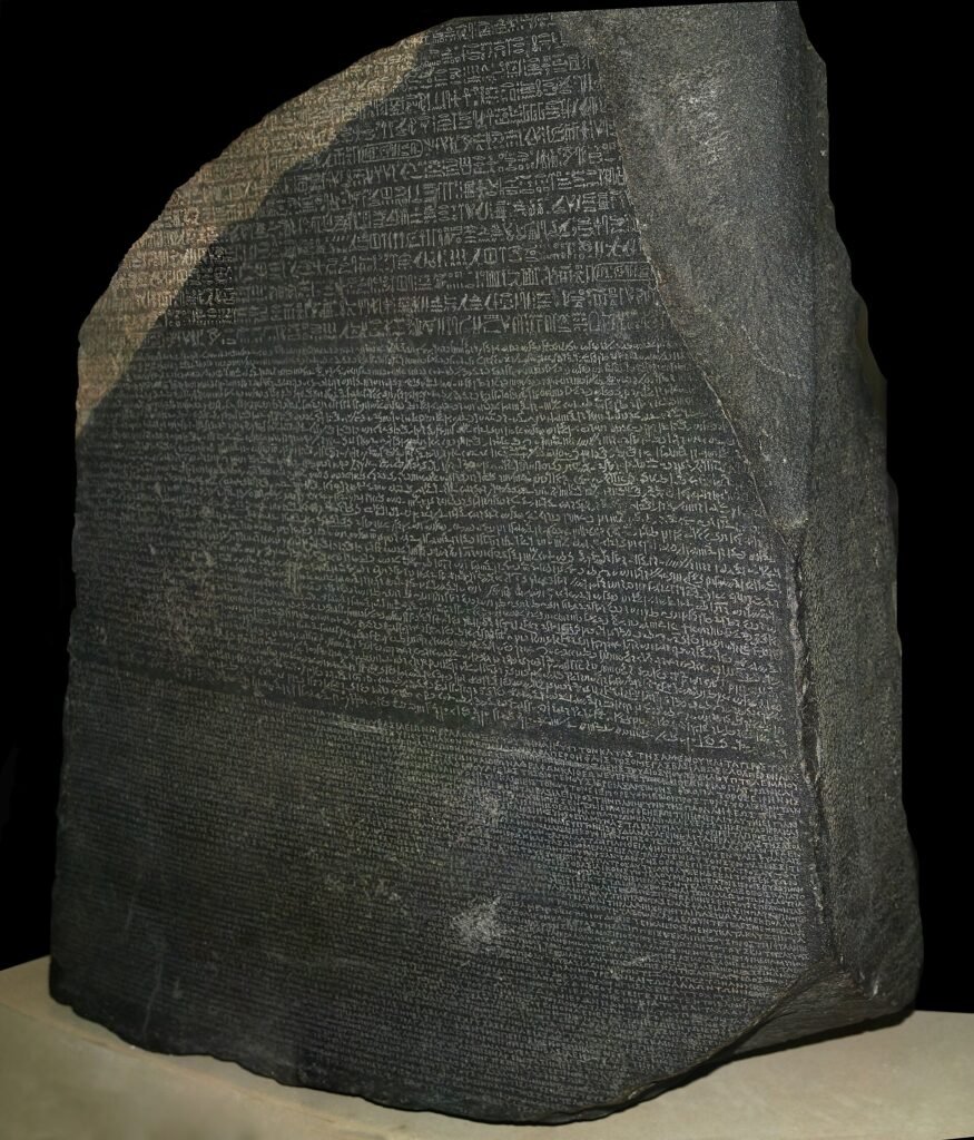 La Piedra de Rosetta de granodiorita oscura sobre pedestal claro y fondo negro, con sus tres escrituras jeroglifica, demotica y griega nitidamente talladas