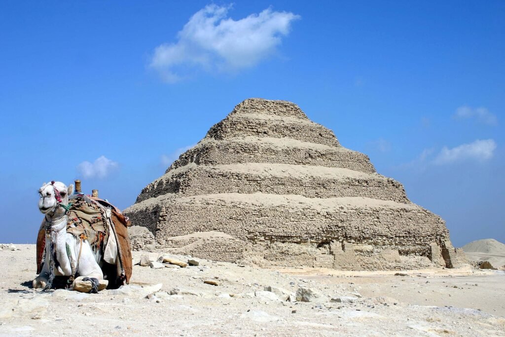 La Piramide Escalonada de Zoser en Saqqara, Egipto, con un camello descansando sobre la arena dorada del desierto bajo un cielo azul intenso, imagen iconica y limpia