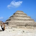 La Piramide Escalonada de Zoser en Saqqara, Egipto, con un camello descansando sobre la arena dorada del desierto bajo un cielo azul intenso, imagen iconica y limpia
