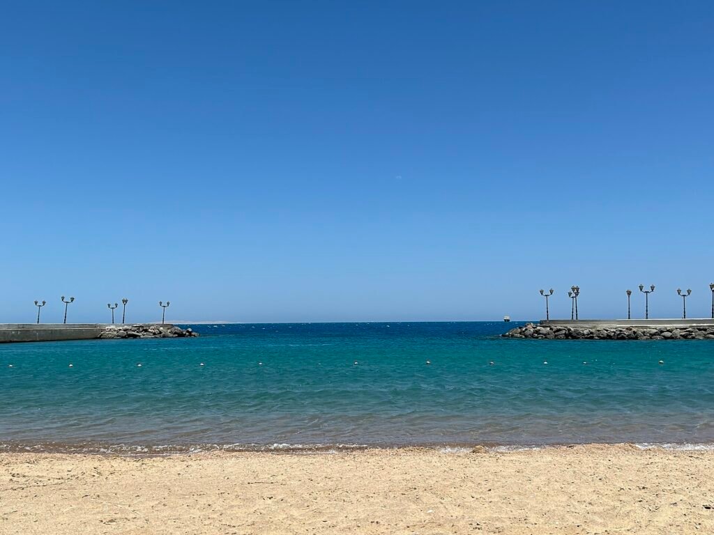 A sheltered Red Sea beach, pale sand in the foreground and turquoise shallow water enclosed by two low rock breakwaters under a cloudless sky