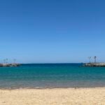 A sheltered Red Sea beach, pale sand in the foreground and turquoise shallow water enclosed by two low rock breakwaters under a cloudless sky