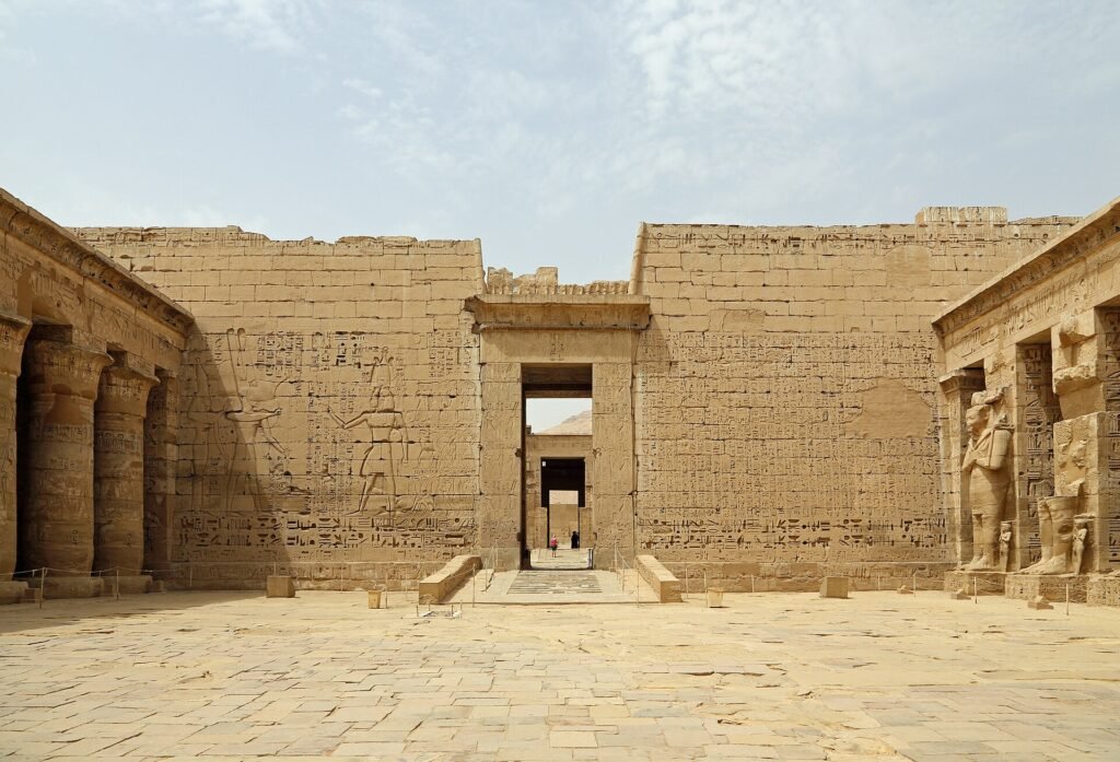 The stone courtyard of Medinet Habu on the Luxor west bank, with tall sandstone walls covered in sunk-relief hieroglyphs and figures of the king, a central doorway opening onto the desert hills beyond, a row of Osiride pillar statues on the right and round columns on the left, under a pale hazy midday sky.