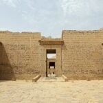 The stone courtyard of Medinet Habu on the Luxor west bank, with tall sandstone walls covered in sunk-relief hieroglyphs and figures of the king, a central doorway opening onto the desert hills beyond, a row of Osiride pillar statues on the right and round columns on the left, under a pale hazy midday sky.