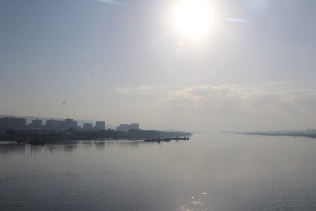 Amplia vista del rio Nilo en el Egipto medio al amanecer, con el sol brillando sobre el agua tranquila y la silueta de una ciudad ribereña a lo lejos