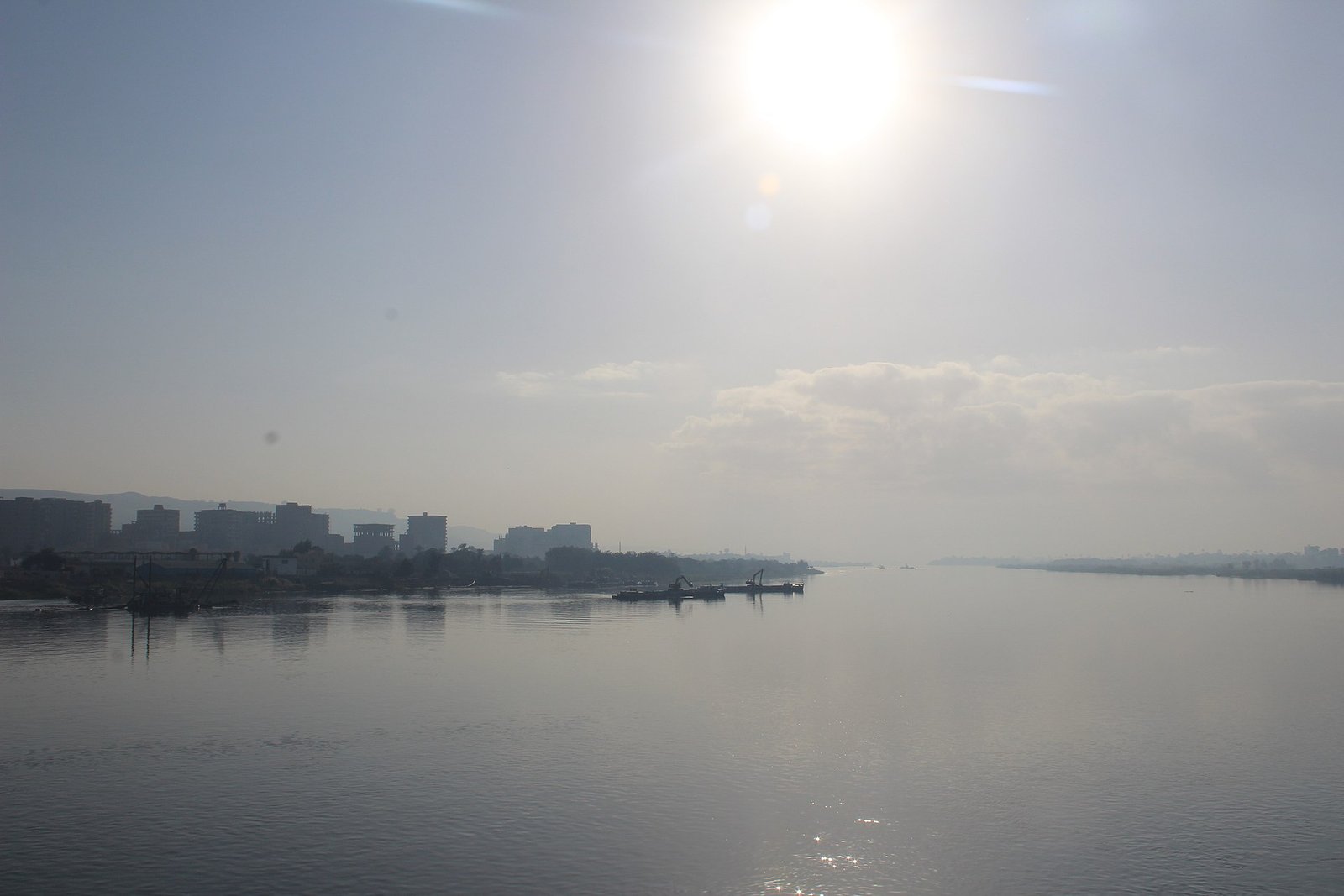 Amplia vista del rio Nilo en el Egipto medio al amanecer, con el sol brillando sobre el agua tranquila y la silueta de una ciudad ribereña a lo lejos